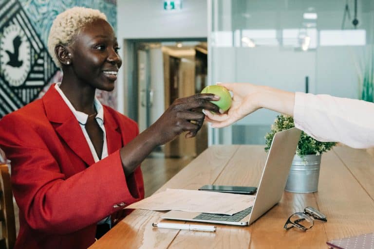 A black female office worker receiving an apple from another woman.