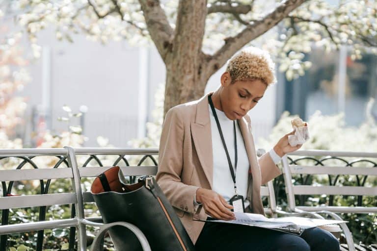 A black American student-worker, sitting on a bench, enjoys a cup of coffee while reading a paper.