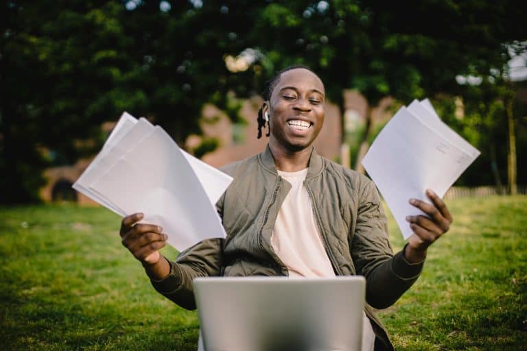 A successful black male student sitting on grass, holding transcripts, with a smile of accomplishment.