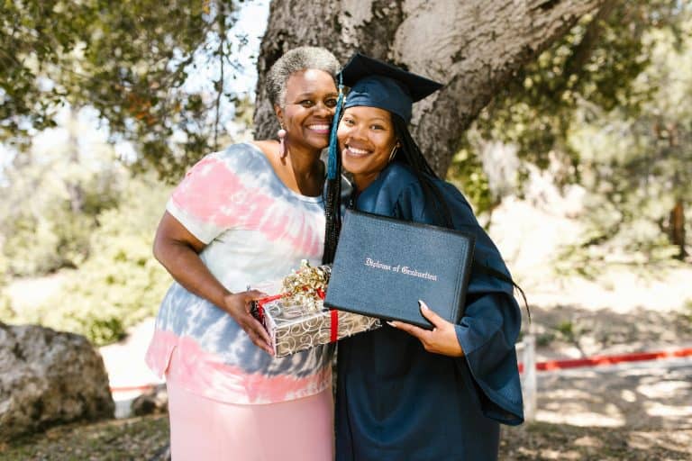 Black American student in graduation gown with her mother, celebrating academic achievement with a gift.